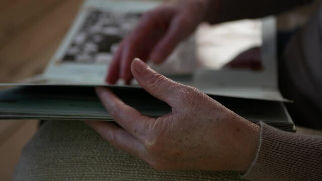 Adult woman looks old photo album at home. Close up view of aged woman's hand with old photo album. Elderly person is leafing through photo album.