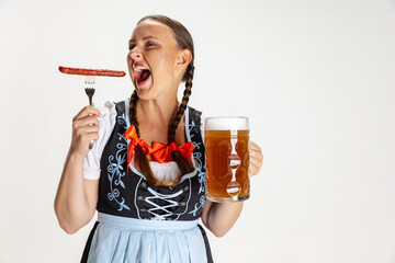 Young beautiful woman, waitress in traditional Austrian or Bavarian costume holding huge beer glass and eating sausage isolated over white background.