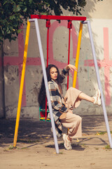 Closeup portrait of young beautiful fashionable woman wearing checkered long coat, beige pants and white blouse . Lady posing on a swing