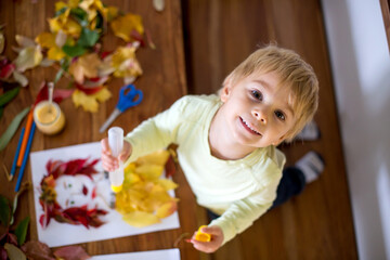 Fototapeta premium Toddler blond child, creating woman portrait of leaves in living room, applying leaves using glue