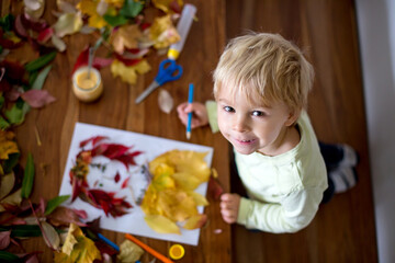 Fototapeta na wymiar Toddler blond child, creating woman portrait of leaves in living room, applying leaves using glue