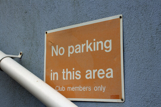 No parking in this area - club members only sign in white letters on an orange background fixed to a cemet wall with a drain pipe below