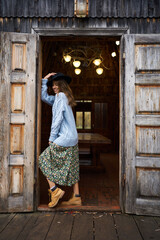 Romantic woman posing with dreamy smile, holding hat, standing in doorway of wooden country house. She is wearing hat, blu sweater and dress