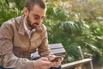 Fototapeta premium A straight faced man sitting on a bench texting