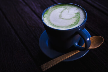 Selective focus on the surface of hot matcha milk tea which have latte art as a flower in the blue beautiful ceramic cup with wooden spoon on the dark brown wooden table background with coyspace