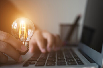 Young businessman using laptop and hand holding glowing light bulb on office desk. Self learning or...