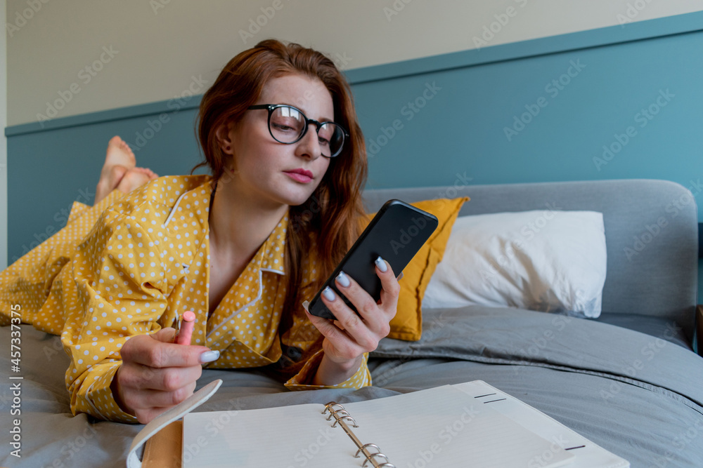 Wall mural young woman in comfortable yellow pajamas works and talks a cell phone while lying the bed in the be