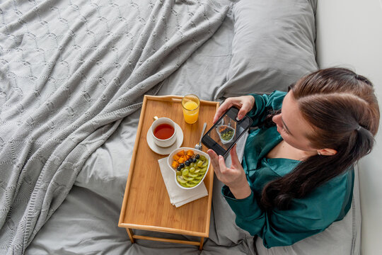 Young woman in having breakfast the morning while lying in bed. Girl takes pictures of her food for social networks. Concept of the use of technology in modern world.