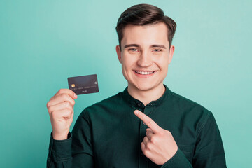 Portrait of reliable guy hold debit plastic card indicate forefinger on teal background