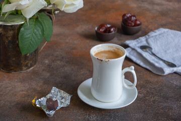 Traces of chocolate on an elegant white cup with a little sipped fresh coffee, a bit of chocolate candy in a golden wrapper next to it, and dates in small bowls in the distance.
