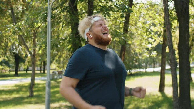 Obese man in in sweaty t-shirt running in park on summer day, then stopping, looking at smartwatch and celebrating new achievement