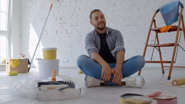 Young caucasian man decorating wall doing house renovation. A male painter taking a break on the floor in front of a painted wall in an apartment. Home improvement concept. New home concept. 4k slow m