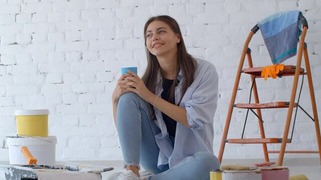 Young caucasian woman decorating wall doing house renovation. A female painter taking a break on the floor in front of a painted wall in an apartment. Home improvement concept. New home concept. 4k sl