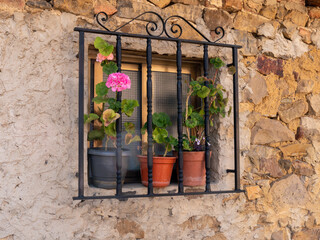 Fototapeta na wymiar A closeup of a window with black bars with sliding glass in a wall of a stone and adobe house and decorated with geraniums in pots
