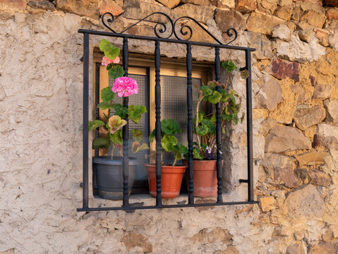A closeup of a window with black bars with sliding glass in a wall of a stone and adobe house and decorated with geraniums in pots