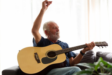 Cheerful senior man playing guitar while sitting on sofa at home. Elderly man learning guitar on...