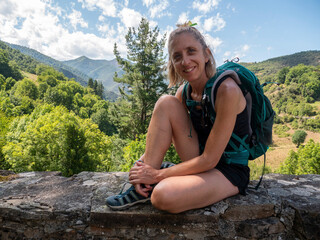 Naklejka na ściany i meble A closeup of a blonde female hiker sitting on a stone wall with a beautiful landscape behind