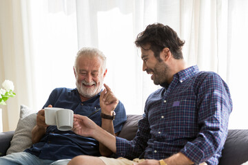 Senior father with adult son talking and drinking coffee while sitting on sofa at home with happy.
