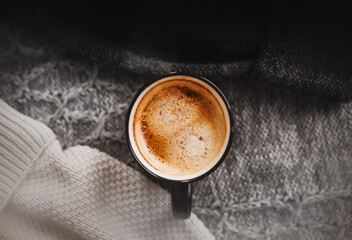 A black mug with a fragrant invigorating latte stands among warm wool sweaters of different colors. The comfort of warm clothes and a hot drink.