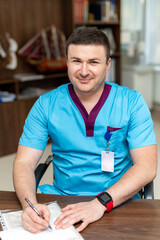 Happy doctor writing something and looking at the camera while sitting at the table at his cabinet. Handsome man in medical uniform. Medicine concept