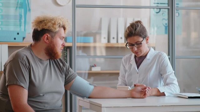 Young Female Doctor Measuring Blood Pressure Of Overweight Male Patient During Health Checkup In Clinic