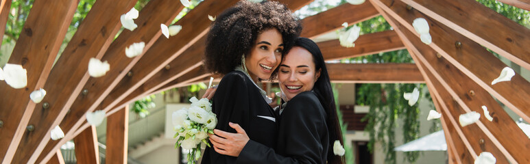 Lesbian african american woman hugging cheerful girlfriend with wedding bouquet under falling petals in park, banner.