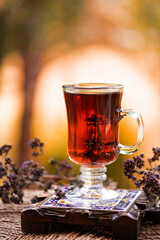 A glass cup of black tea with herbs on the handcrafted wooden and ceramic base with an autumn tree with bokeh on the background