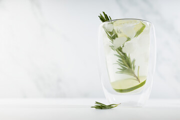Organic mojito cocktail with citrus fruit slices, ice cubes, rosemary twig in transparent mug on white wood table, soft light marble wall.