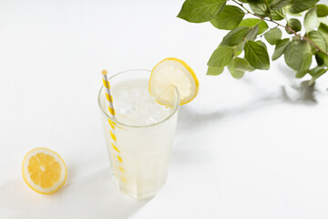 Fresh lemon tonic drink of organic lemons in misted glass with ice cubes, straw, yellow lemons on white wood table in sunny summer day.
