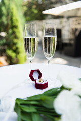 Wedding rings near glasses of champagne and blurred flowers outdoors.