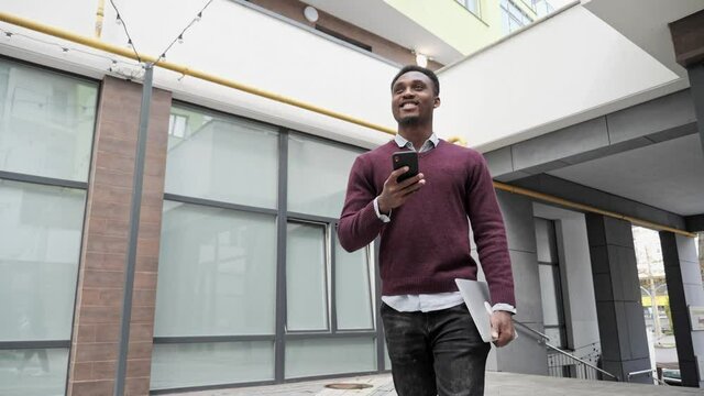 Dark-skinned Handsome Man having a Slow walk in the Town, enjoying Conversation. Typing Messages on his Smartphone and Holding Laptop. Wearing stylish clothes. Having nice Hairstyle. Modern Technology - Powered by Adobe