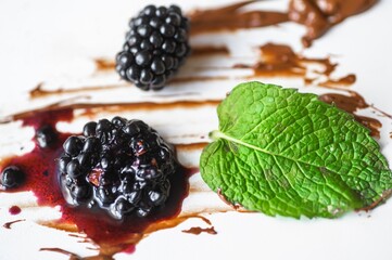 2 blackberry, one is crushed with sauce, green leaf on dirty white background with chocolate fleck.