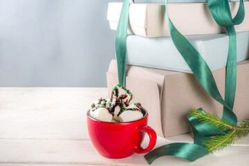 Big stack of Christmas gifts with hot chocolate, stay on it like pedestal, cozy pastel colored craft paper gift boxes with green festive Xmas ribbon, cup of cocoa with marshmallow, copy space