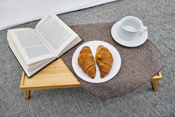 A clean white mug, a book, a croissant in a cozy home decor. Food, books on a wooden tray. Breakfast on the bed in a bright room. The concept of enjoying the seasons, winter mood, reading, breakfast