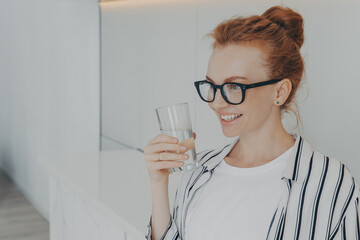 Smiling redhead young woman holds glass of water drinks aqua for body refreshment