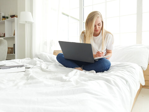 blond young female making notes with pen with latop on her nees in bed
