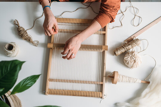 Hands of woman weaving wool rug at the table with flowers and threads