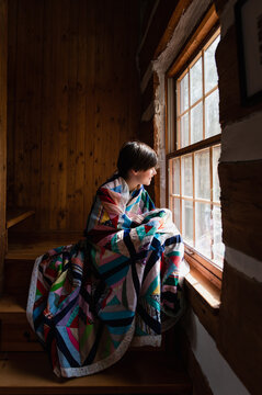 Young boy wrapped in a quilt looking out the window of a log cabin.