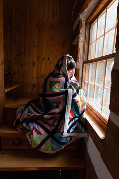 Young Boy Wrapped In A Quilt Looking Out The Window Of A Log Cabin.