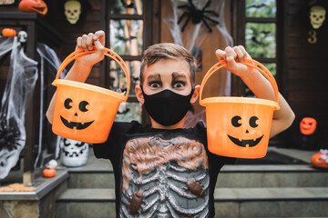 boy in skeleton costume and black medical mask holding halloween buckets near blurred cottage porch