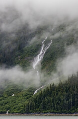 Thin ribbon like waterfalls run down tall mountains surrounded by mist in Alaska.