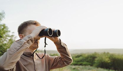Fototapeta na wymiar A young man looks through binoculars at a beautiful hilly landscape.The concept of hunting, travel and outdoor recreation. Banner with copy space.A traveler or hunter is observing through binoculars.