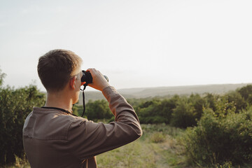 A young man looks through binoculars at a beautiful hilly landscape.The concept of hunting, travel and outdoor recreation. Banner with copy space.A traveler or hunter is observing through binoculars.