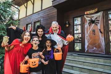 family in creepy costumes holding halloween attributes and shouting near house