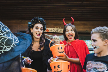 woman with candies near excited kids holding buckets and shouting in creepy halloween costumes