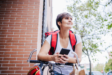 Fototapeta na wymiar young rider woman wearing red backpack holding paperboard box of food in city. Delivery service concept. Woman checking order in mobile phone
