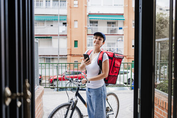 Young rider woman wearing red backpack delivering food on a bike, checking order with smart phone...