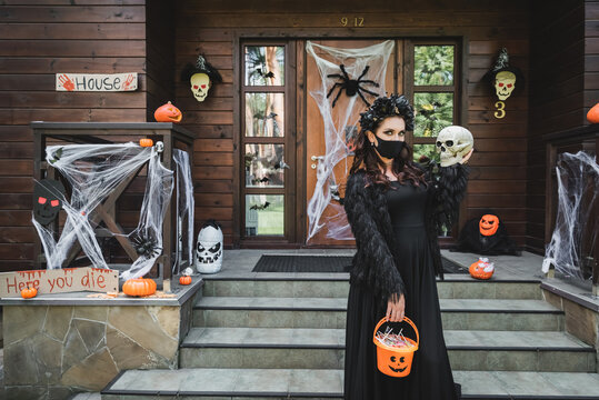 woman in halloween costume and black medical mask standing with skull and bucket of sweets near decorated porch