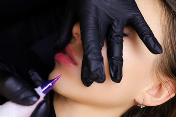 the master of permanent makeup holds his lips with his fingers and performs permanent makeup with a...