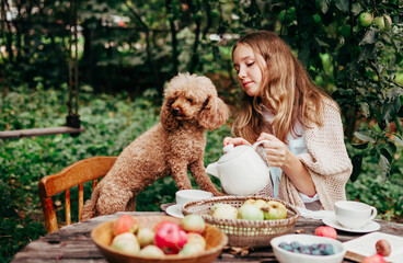 teenage cute girl with her poodle dog sits on wooden table set for tea in the garden under an apple tree, cozy and comfortable autumn still life
